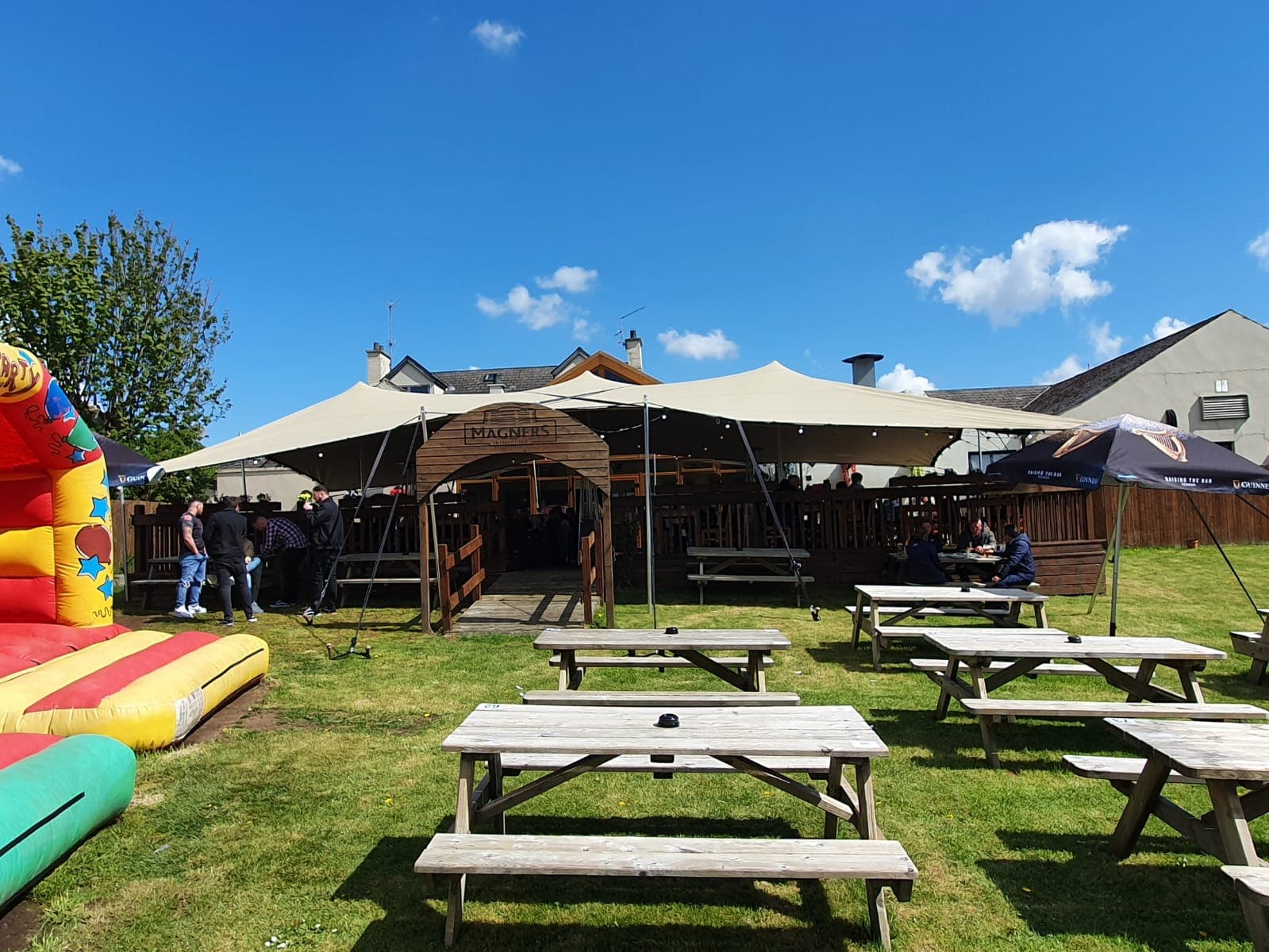 Outdoor bar terrace covered by a cream stretch tent on a sunny day, with picnic benches on the grass