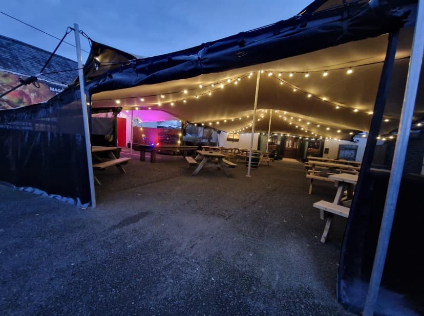 Evening view inside a stretch tent with warm festoon lighting and picnic benches in an outdoor hospitality courtyard