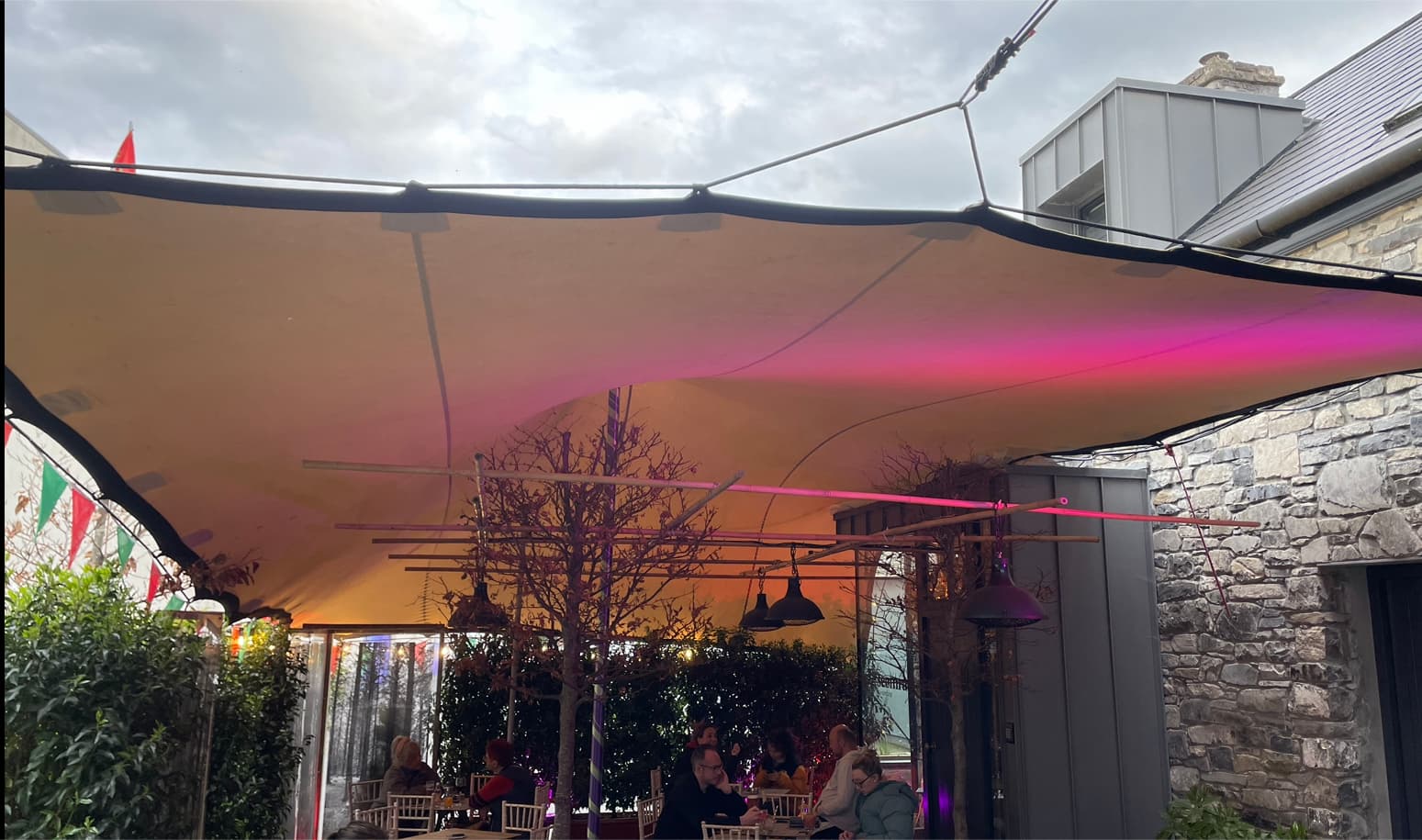 Stretch tent canopy covering an outdoor courtyard seating area beside a stone building, lit with pink accent lighting at dusk
