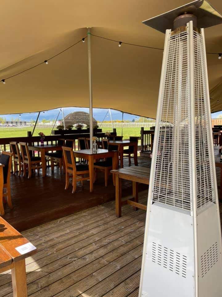 Outdoor dining setup beneath a stretch tent with wooden tables, string lights and a patio heater