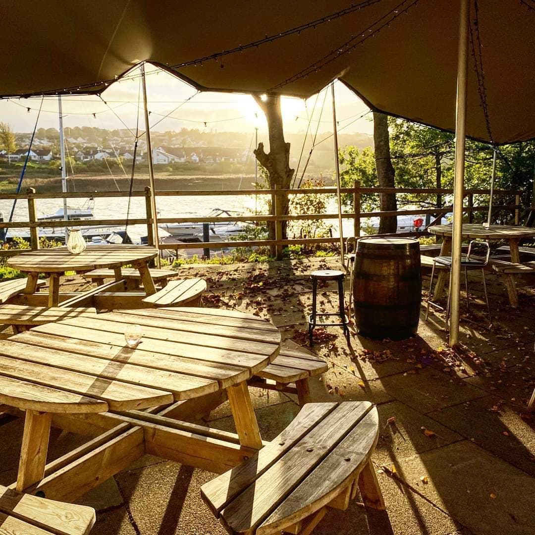 Riverside outdoor seating area beneath a stretch tent at golden hour with picnic tables and warm string lighting