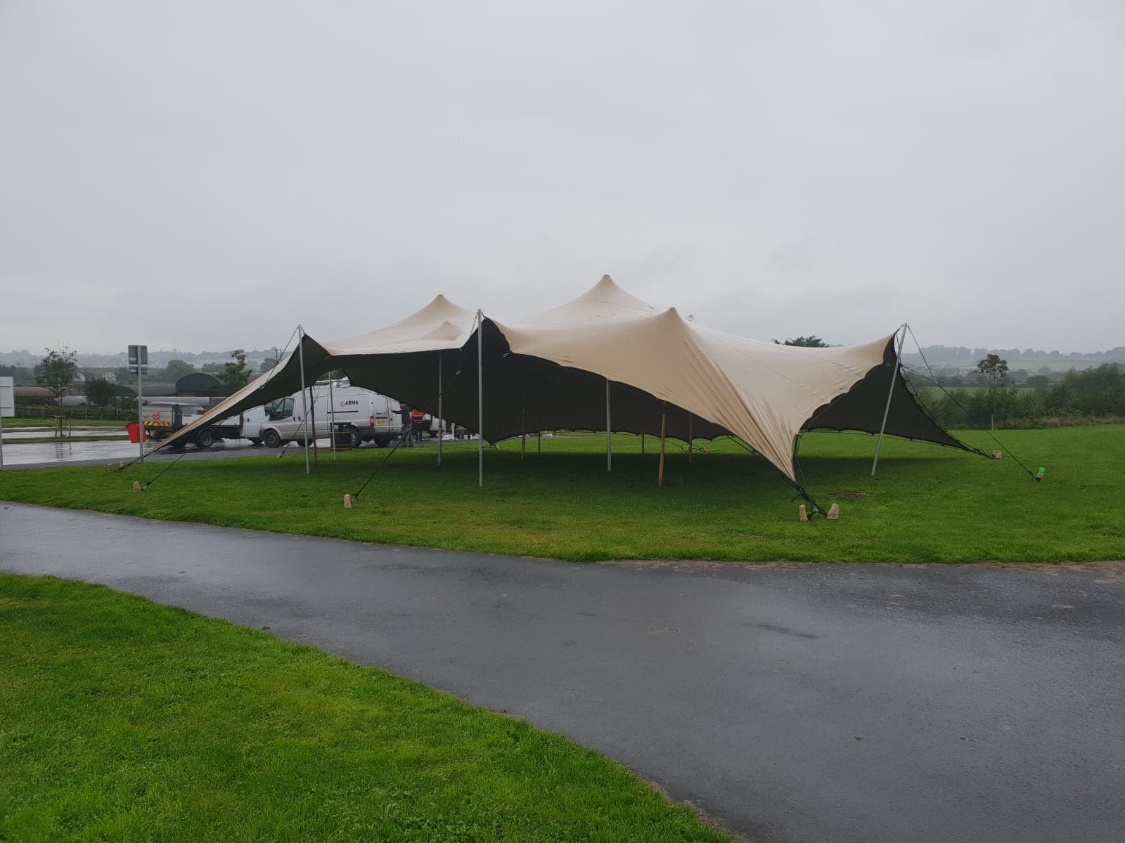 Beige stretch tent set up on a grassy field beside a wet roadway under an overcast sky