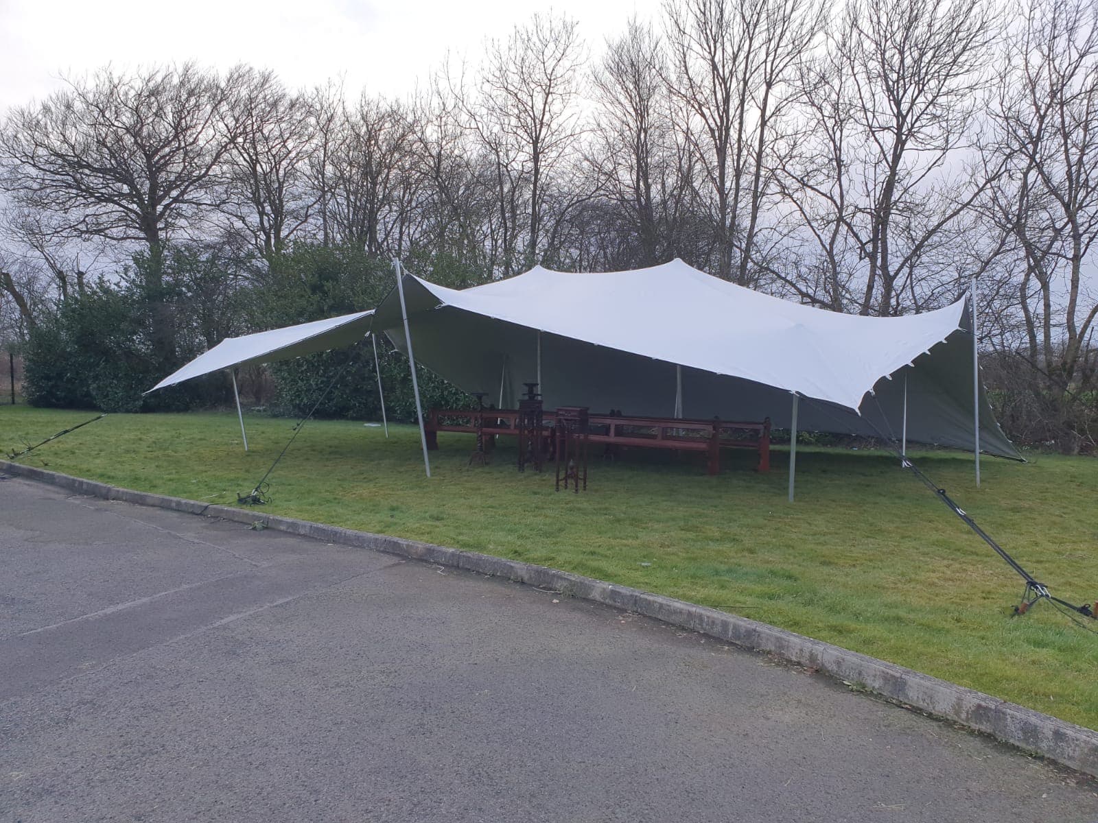 White stretch tent set up on a grass verge beside a paved area with bench seating and bare trees in the background