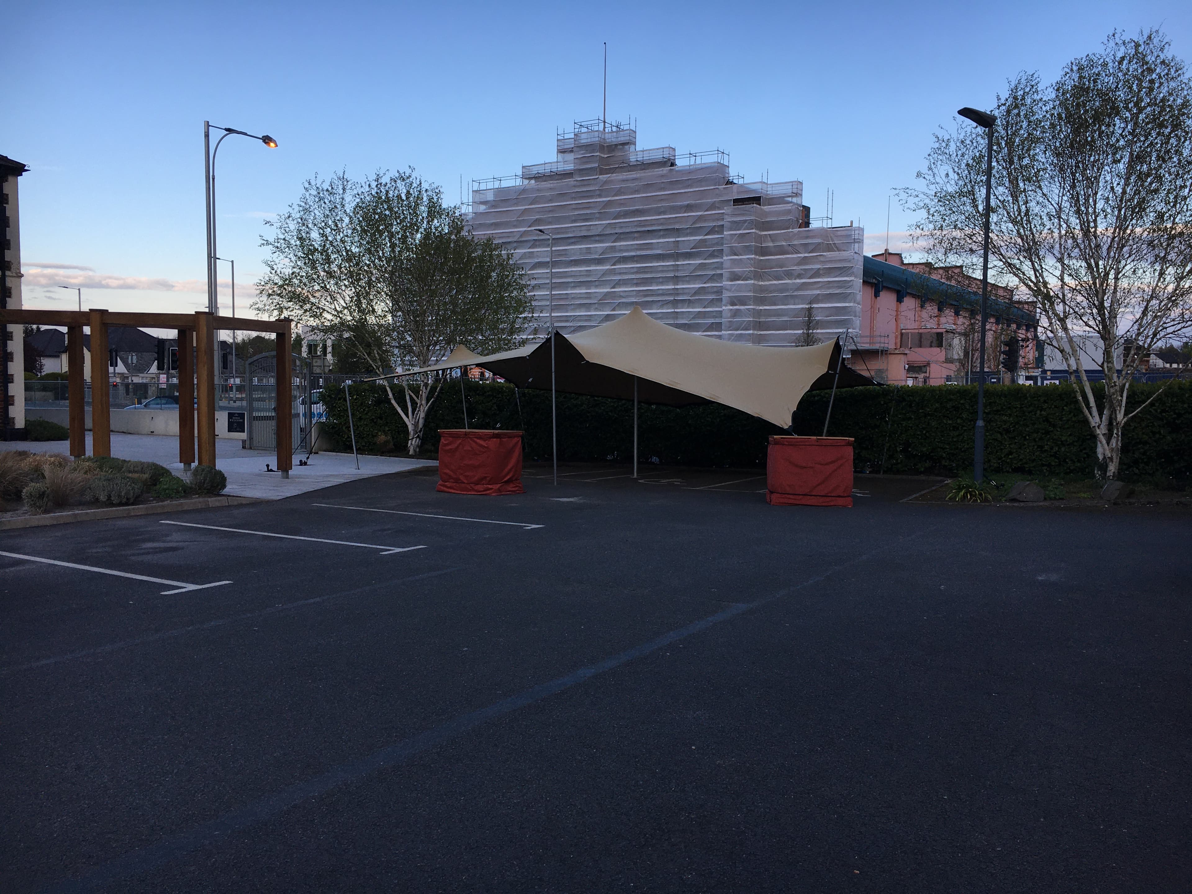 Stretch tent set up in a car park at dusk beside trees and an urban building