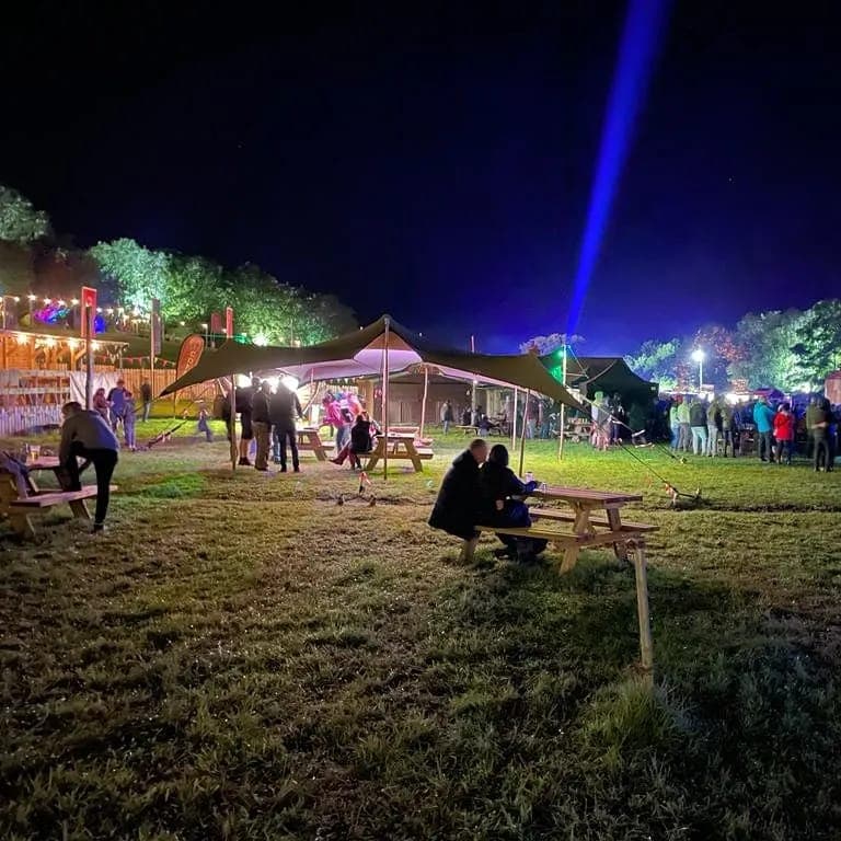 Nighttime festival scene with a stretch tent over picnic benches and groups of people on a grassy field