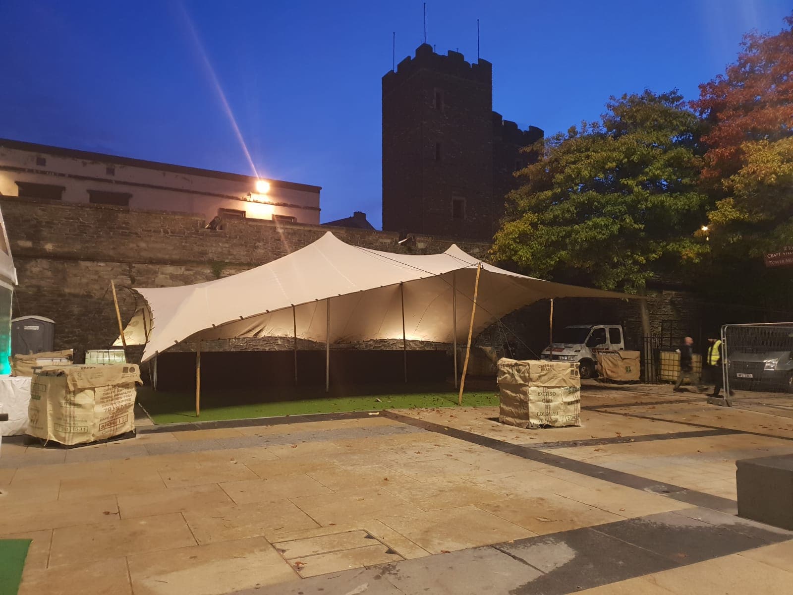 Illuminated stretch tent set up in a historic courtyard at dusk with a stone tower and trees in the background
