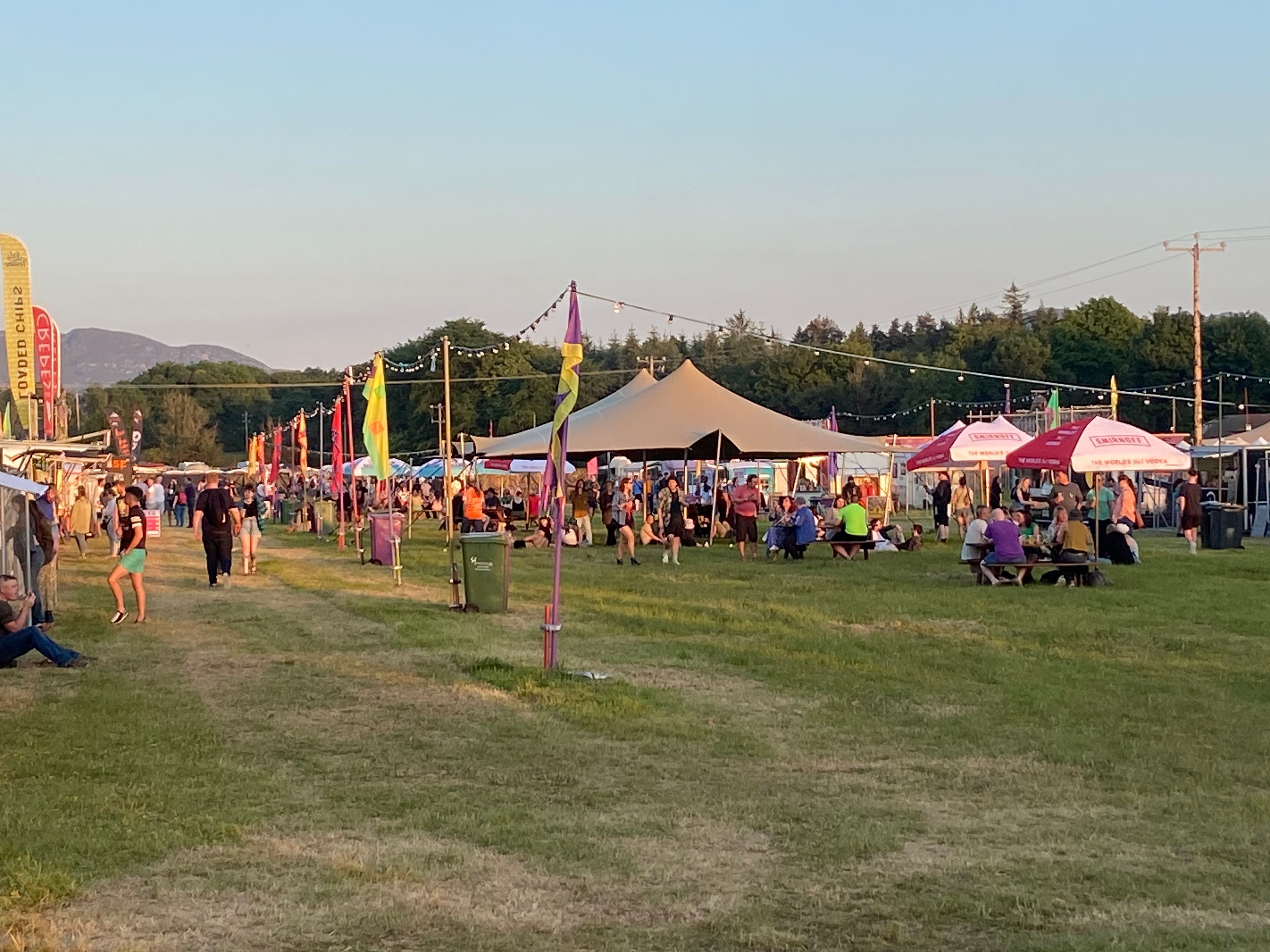 Outdoor festival scene with a central stretch tent, people relaxing on the grass, vendor stalls, and string lights at dusk