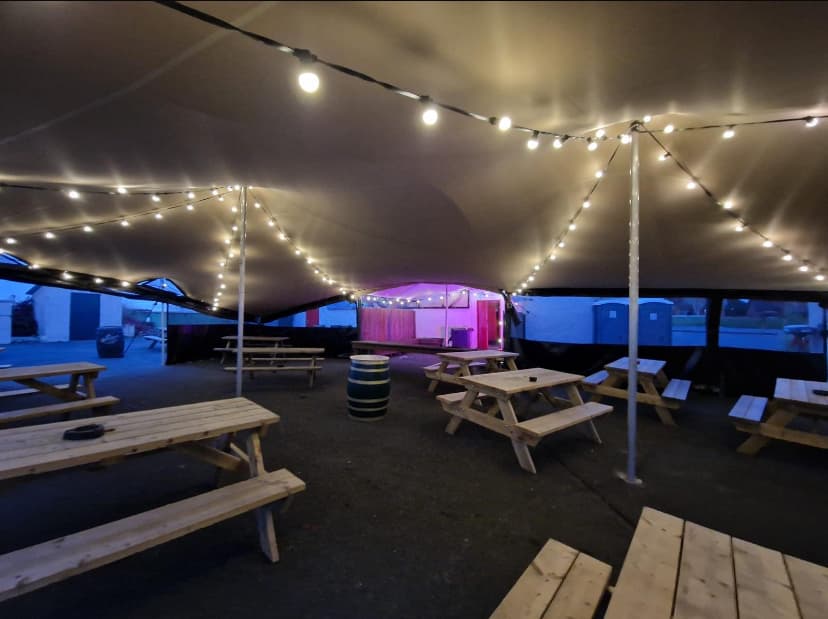Interior of a stretch tent with festoon lighting and picnic bench seating at dusk