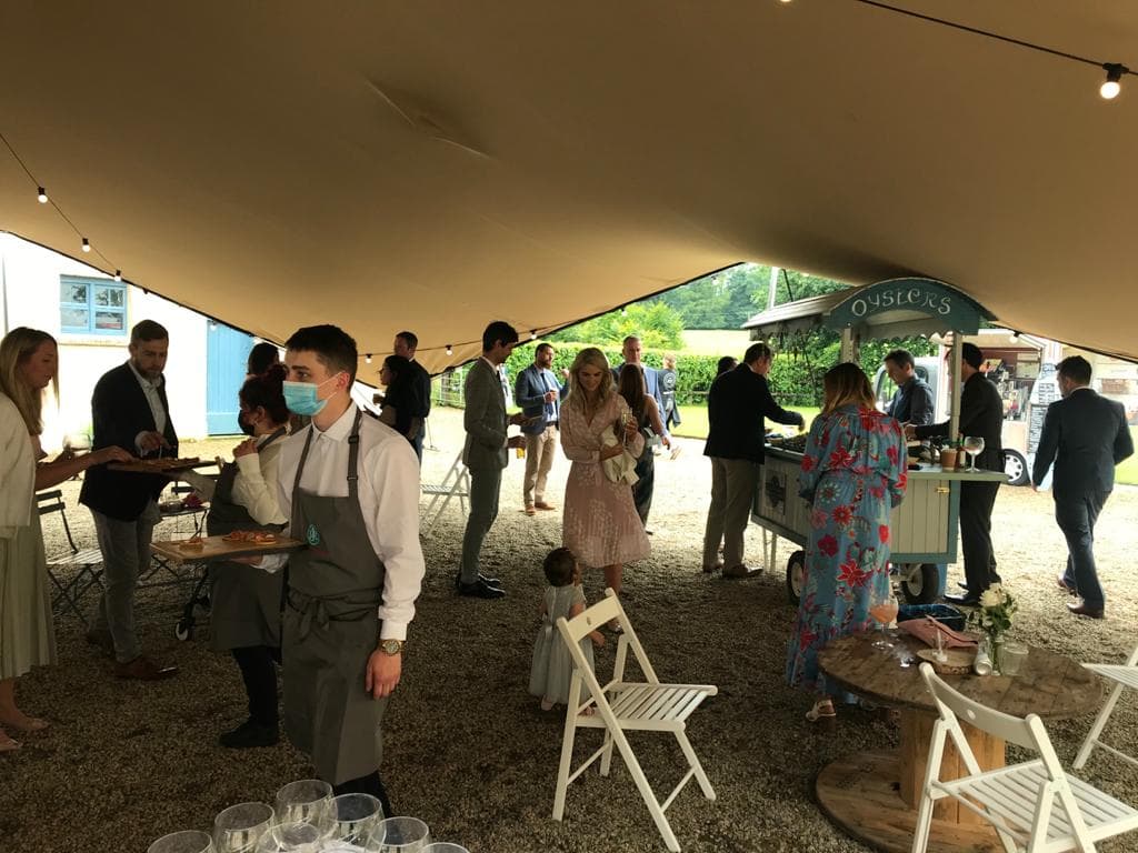 Guests mingling under a stretch tent at an outdoor catered event with servers and a food cart