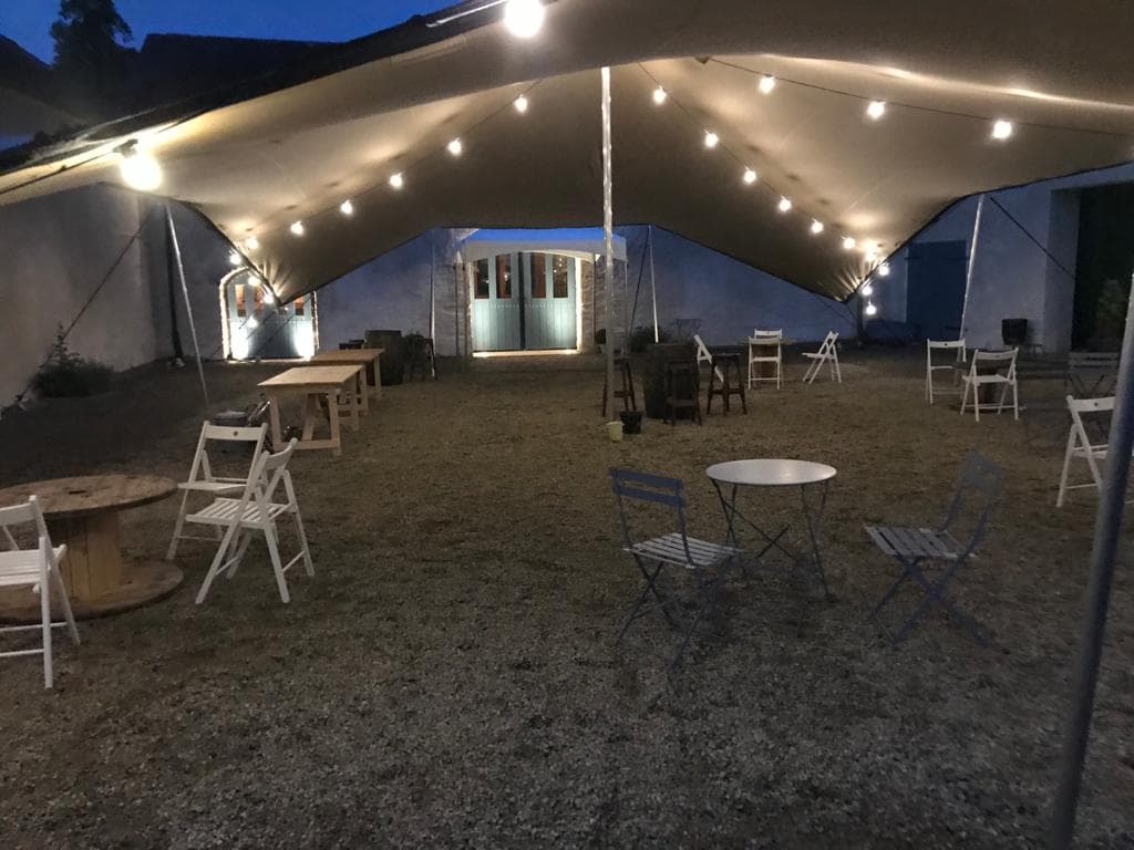 Evening view beneath a stretch tent with festoon lighting, scattered chairs and small tables in a courtyard setting