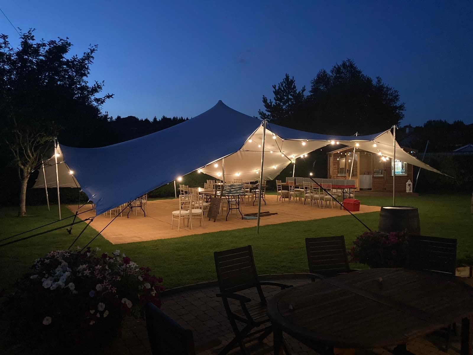 Illuminated stretch tent set up in a garden at dusk with string lights, tables and chairs underneath