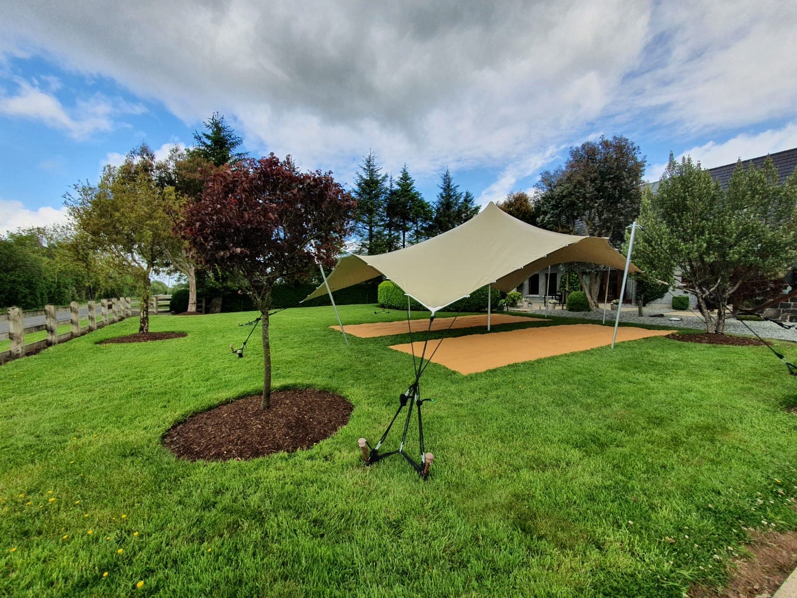 Stretch tent set up on a manicured lawn beside a house, with trees surrounding the garden under a dramatic cloudy sky