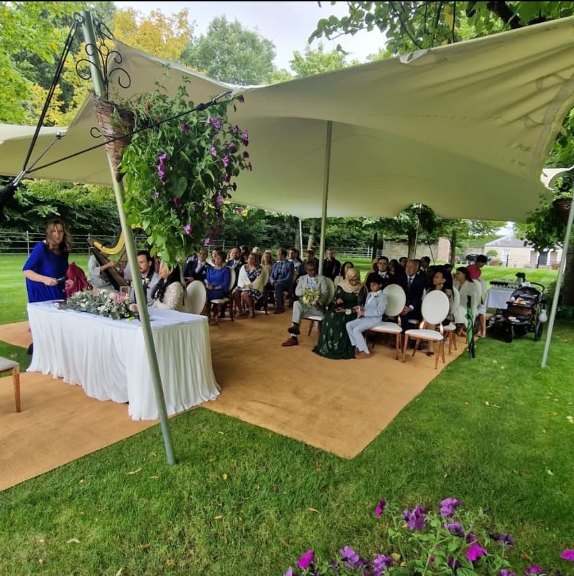 Outdoor wedding ceremony under a cream stretch tent in a garden with seated guests and floral decor