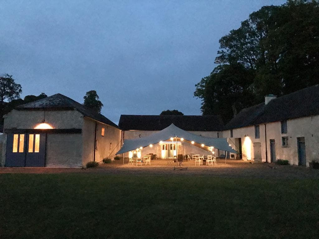 Stretch tent lit with warm festoon lights in a courtyard between rustic buildings at dusk
