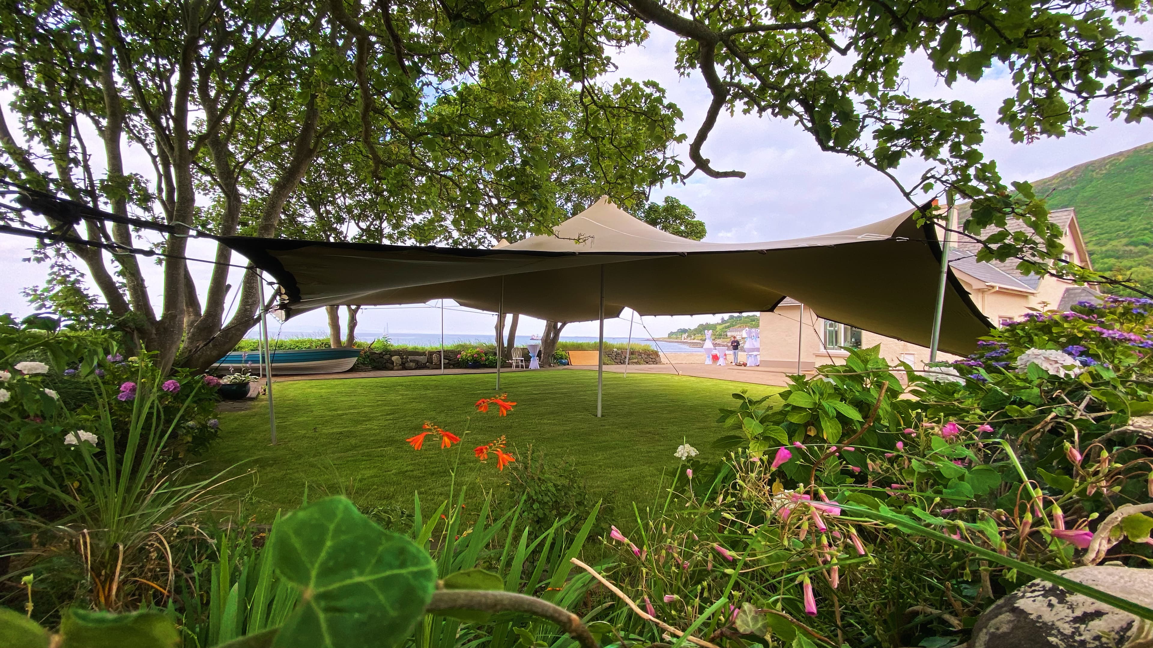 Stretch tent set up on a landscaped seaside garden lawn beneath mature trees beside a house
