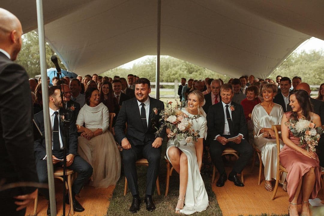 Wedding ceremony guests smiling beneath a stretch tent as the bride and groom sit at the front