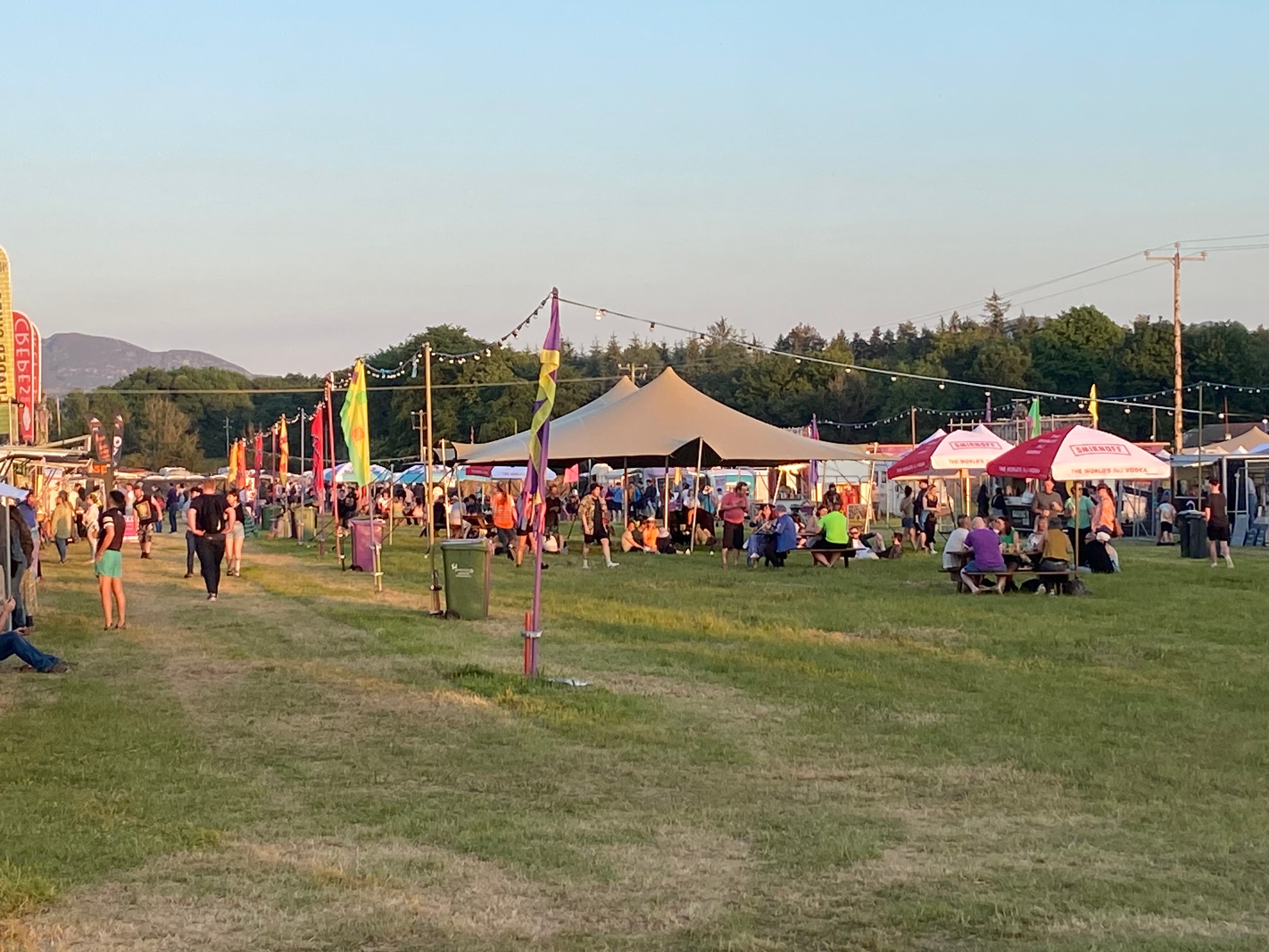 Stretch tent at a busy outdoor festival with people gathering on a grassy field at sunset